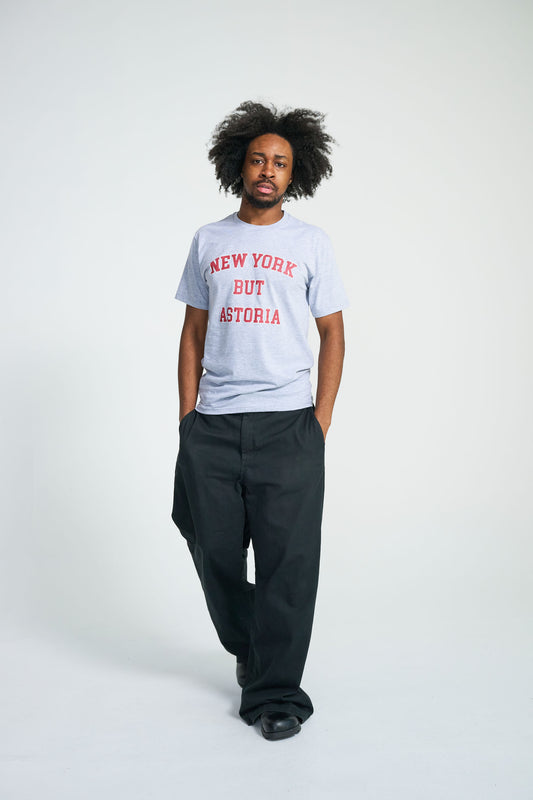Person wearing a New York But Astoria light gray t-shirt with text and dark pants on a white background