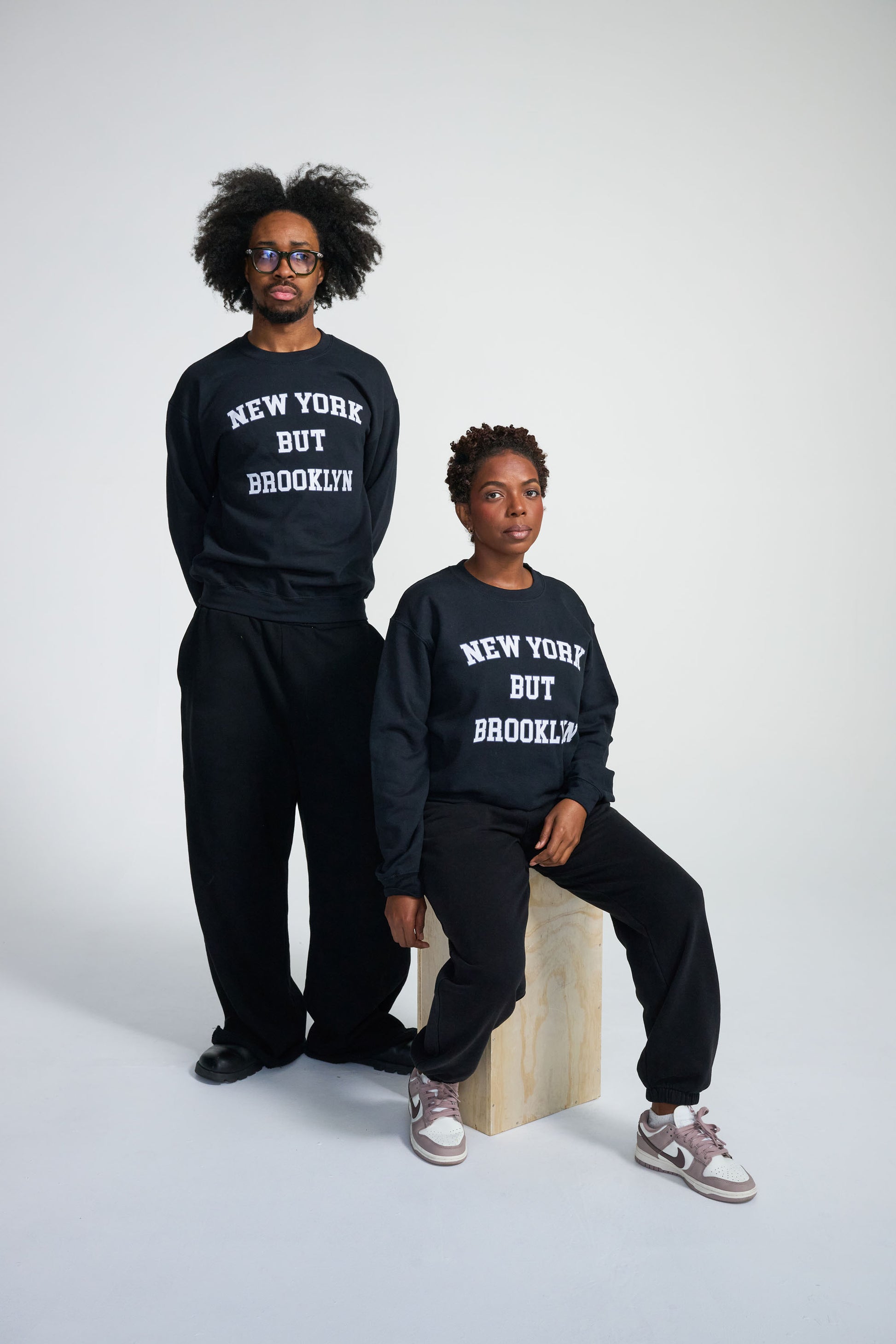 Two people wearing matching black sweatshirts with 'New York But Brooklyn' text on a white background.