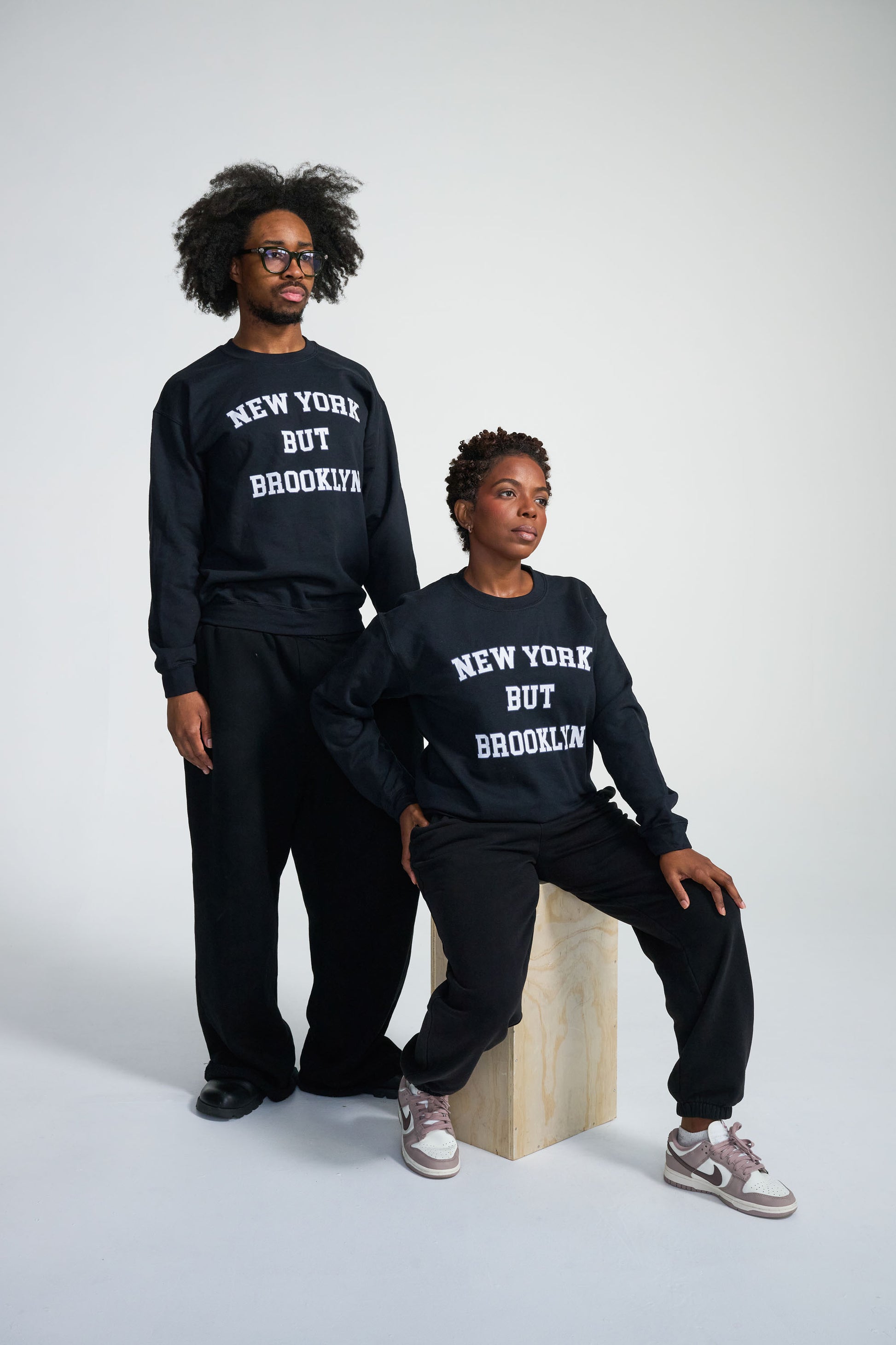 Two people wearing matching sweatshirts with 'New York But Brooklyn' text on a white background